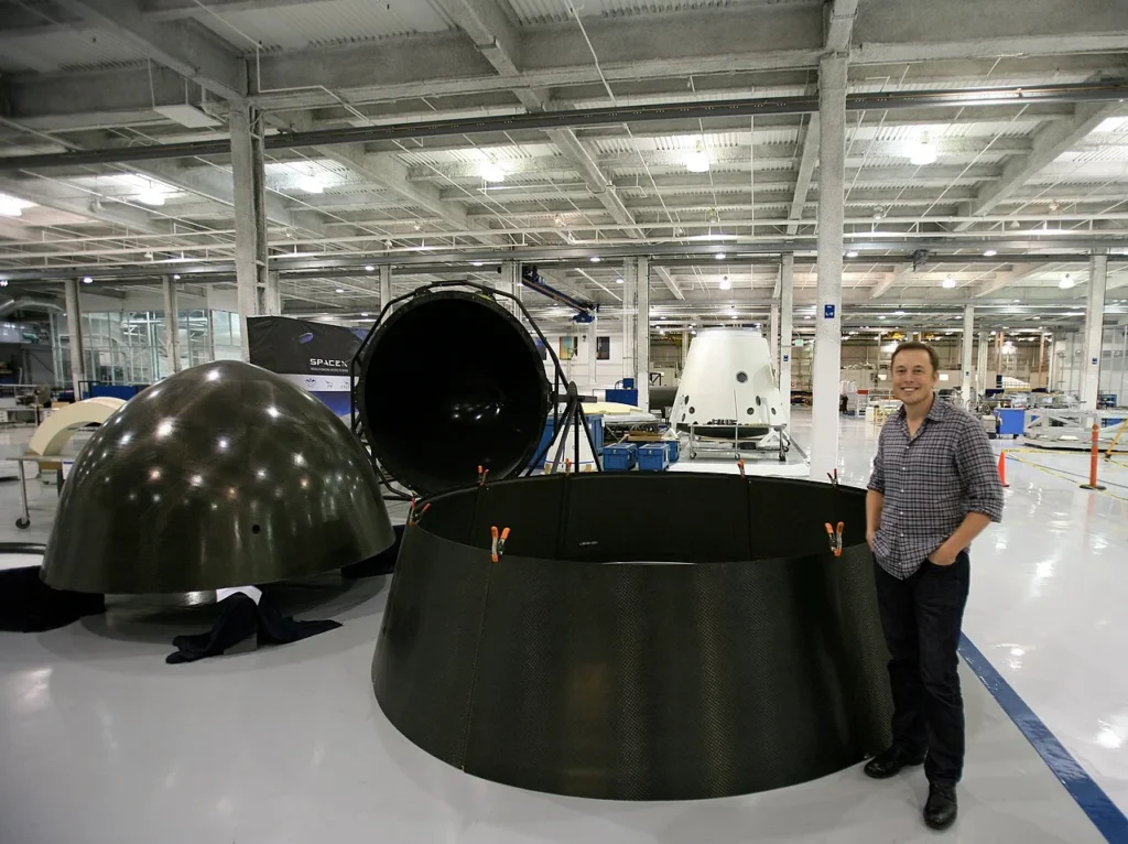 Elon Musk standing next to carbon-fiber spacecraft components inside a SpaceX factory hangar.