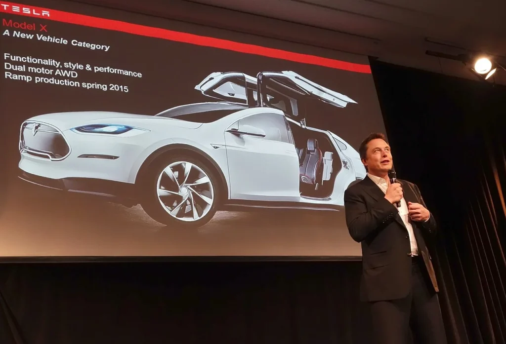 Elon Musk speaking into a microphone on stage at a Tesla shareholder meeting in front of a Model X presentation screen.