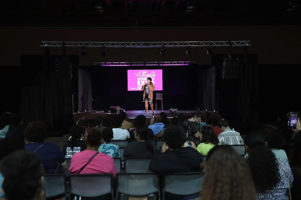 Kim Fields standing on stage speaking at the Ultimate Women's Expo in front of a seated audience.