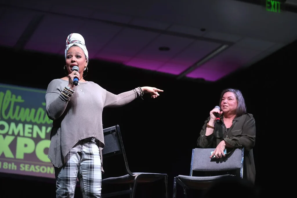 Kim Fields holding a microphone and speaking on stage next to her sitting co-star Mindy Cohn at a public event.