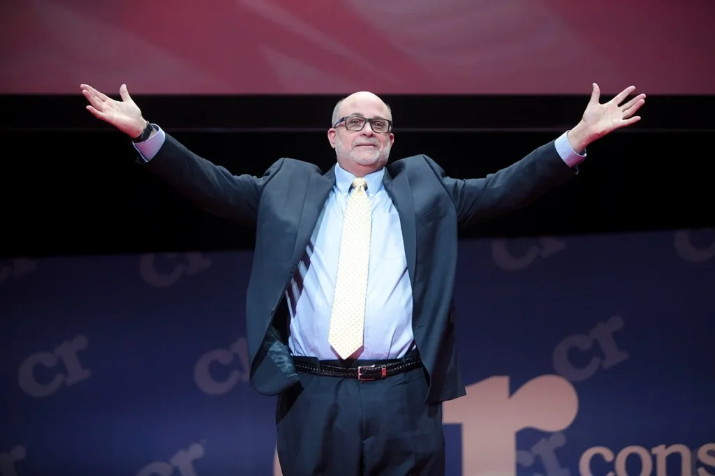 Mark Levin standing on a stage with arms spread wide smiling.