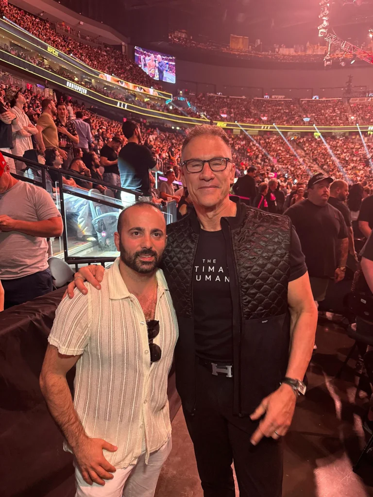 Gary Brecka in a black vest and Artur Shakhnazarov posing for a photo inside a crowded UFC arena during a fight event.