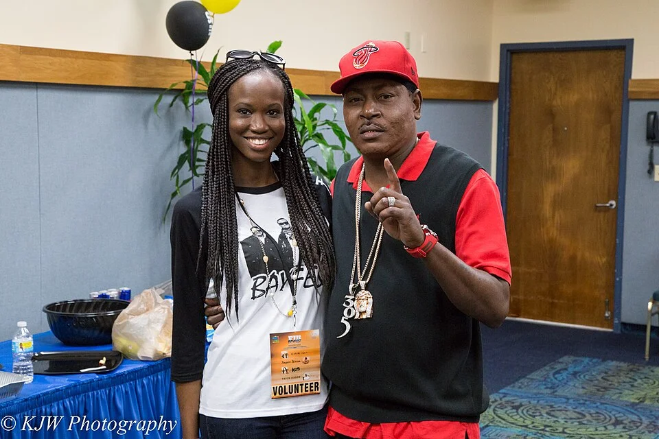 Rapper Trick Daddy posing for a photo with a volunteer at the Bayfest 2015 event, wearing a red Miami Heat hat and heavy gold chains.