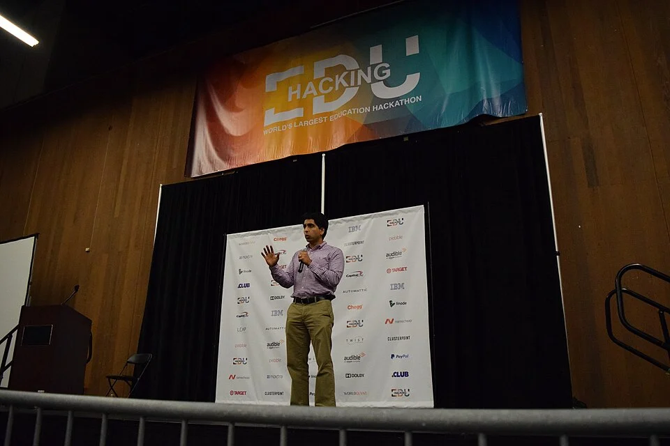 Sal Khan speaking on stage at the Hacking EDU conference in front of a sponsor banner.