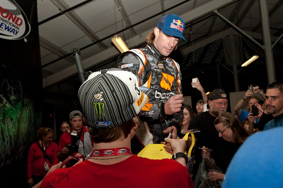 Travis Pastrana in a Red Bull cap and racing suit signing autographs for fans