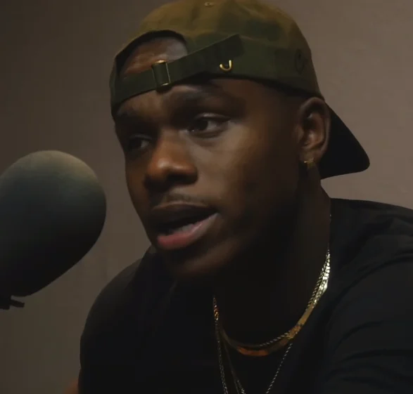 Rapper DaBaby speaking into a studio microphone wearing a green cap and gold chains.
