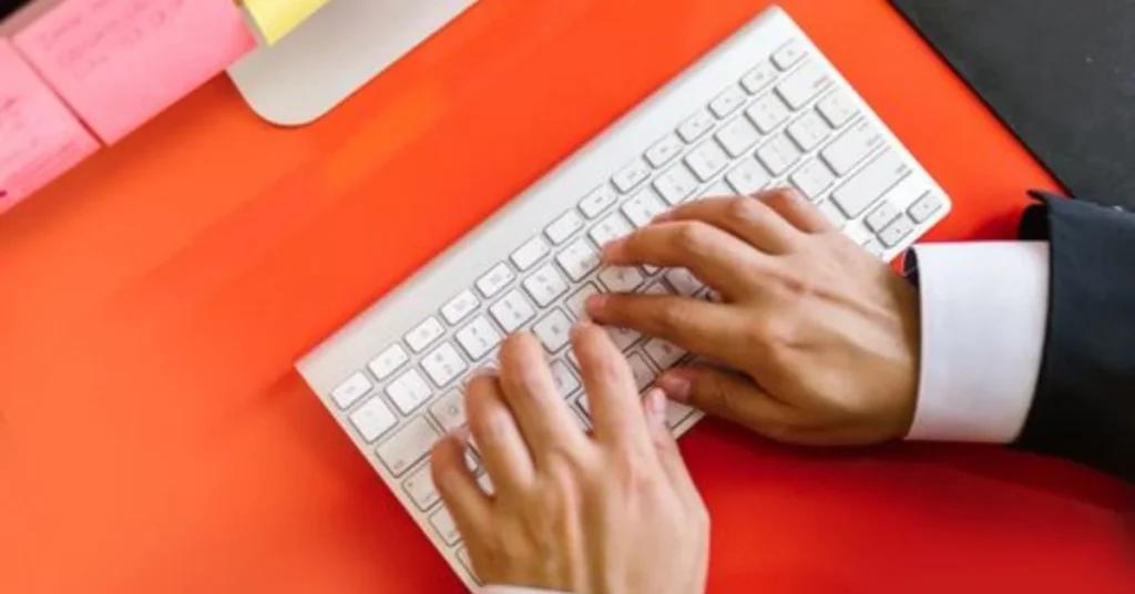 Overhead view of a person in a suit typing on a slim white wireless keyboard on a bright red desk with sticky notes